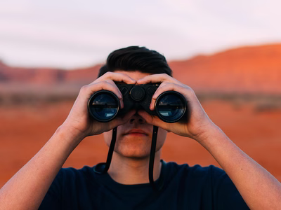 A man looks in to the camera through a pair of binoculars.