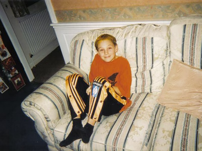 Darren as a young man (circa 10 years old) sat on a white sofa with stripes, wearing an orange t-shirt and black and orange tracksuit bottoms.