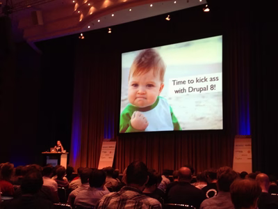A humourous keynote slide from a DrupalCon. The slide shows a toddler clenching a fist and some wording saying 'Time to kick ass with Drupal 8'.