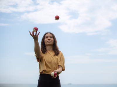 A woman standing trying to juggle, she’s holding two balls and is looking at the third ball in the air.