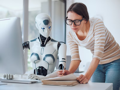 A robot at an office desk whose work is being reviewed by a female colleague who looks less than impressed.