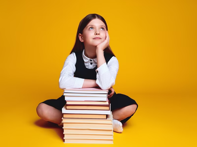 A young girl seating on the floor, leaning into a stack of books daydreaming with one hand on her face.