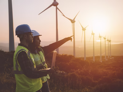 Two young electrical engineers (a woman and a man) standing in front of wind turbines checking and working out technical problems and logging the results of measurements on a laptop.