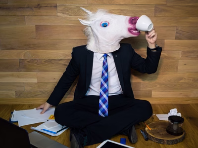 Man seating on the floor wearing a suit and an unicorn head custom. He is trying to drink from a white cup and has laptop and some paper on the floor next to him. 