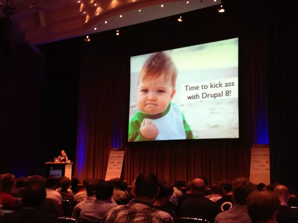 A humourous keynote slide from a DrupalCon. The slide shows a toddler clenching a fist and some wording saying 'Time to kick ass with Drupal 8'