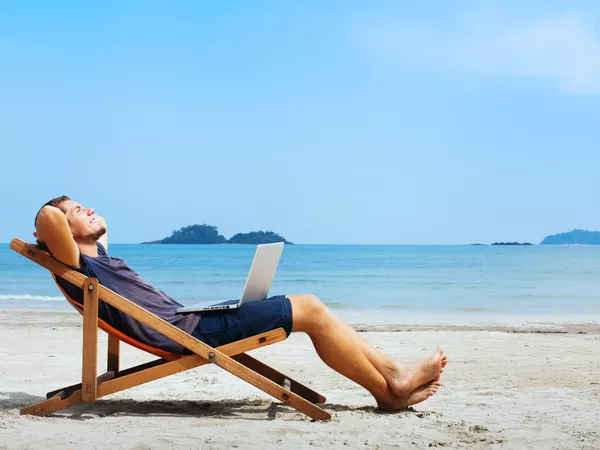 A man in shorts and t-shirt lying back in a deckchair on a white sandy beach with a laptop on his lap.