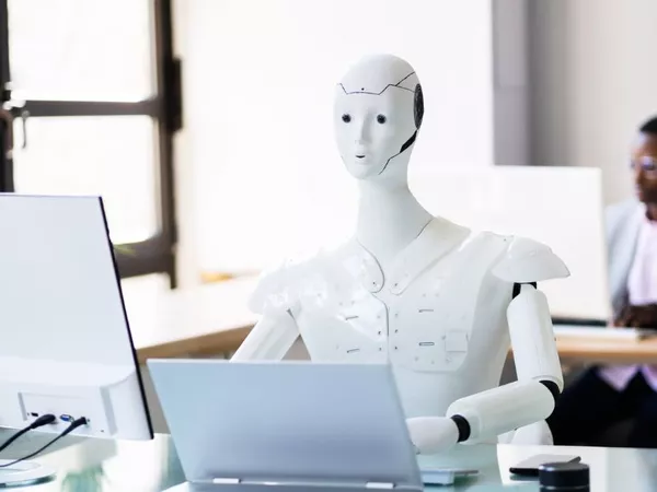 A robot at an office desk working with a laptop and another man working in desk behind him using a computer. 