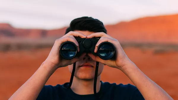 A man looks in to the camera through a pair of binoculars.