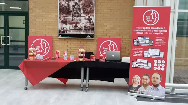 The Real Life Digital stand before the crowds arrived - a long table with giveaways and cupcakes on it. In the background are two large Real Life Digital logos printed on to canvas and to the right is a pop up banner featuring some of Real Life Digital&apos;s work.