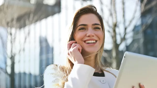 A blonde woman smiles as she walks holding an iPad and a phone to her ear.