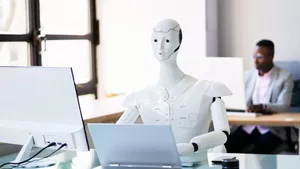 A robot at an office desk working with a laptop and another man working in desk behind him using a computer.