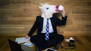 Man seating on the floor wearing a suit and an unicorn head custom. He is trying to drink from a white cup and has laptop and some paper on the floor next to him.